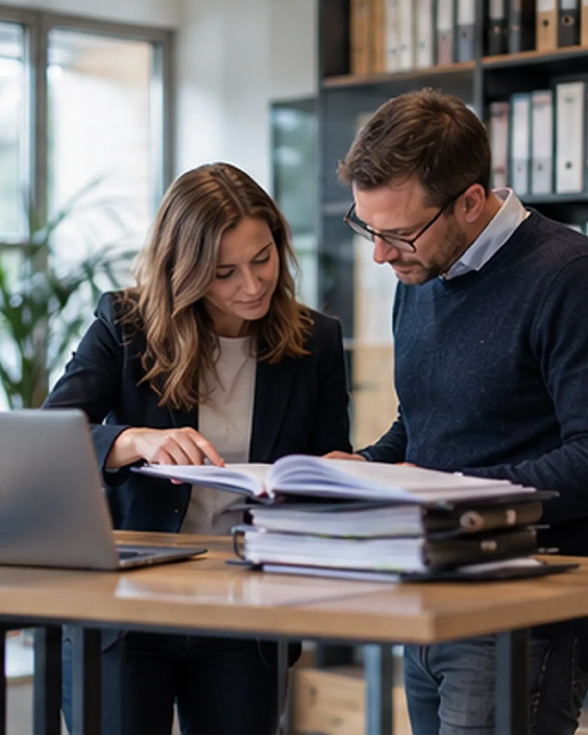 Twee collega's bekijken samen een dossiermap naast een laptop in een kantoor met archiefkasten.