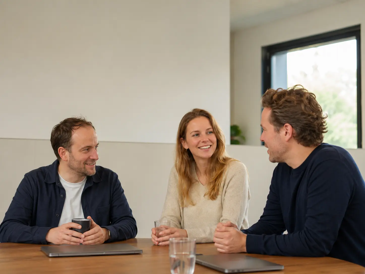 Drie consultants voeren een informeel gesprek aan tafel in een lichte werkruimte.