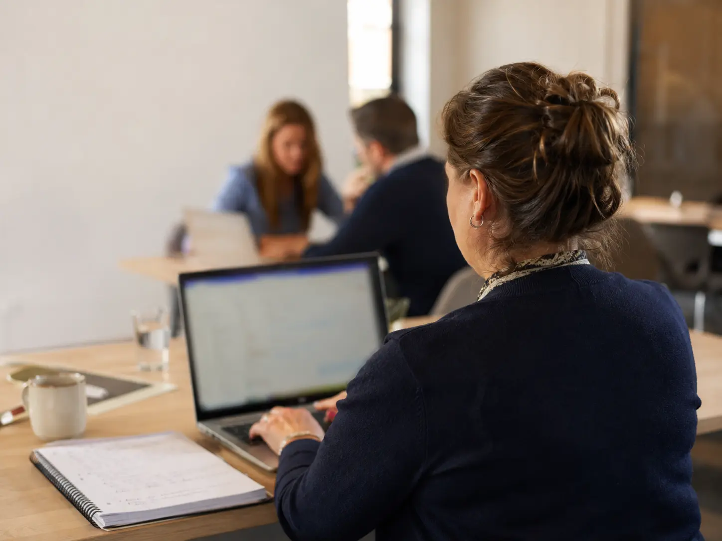 Deelnemer typt geconcentreerd op haar laptop met een notitieblok naast zich en overleggende collega's op de achtergrond.